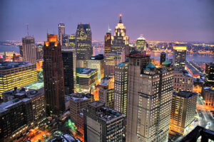 Nighttime cityscape of Detroit, where outdoor lighting enhances the illuminated skyscrapers and buildings, with the Detroit River gracefully flowing in the background.
