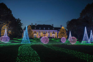 A large mansion is illuminated at night with decorative lights, including glowing cones, spheres, and green paths, set in a landscaped garden.