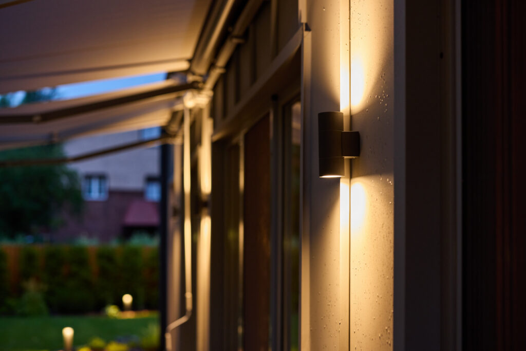 Modern outdoor wall sconce mounted beside a glass door, casting warm ambient light along a home’s exterior at dusk with a softly lit garden in the background.